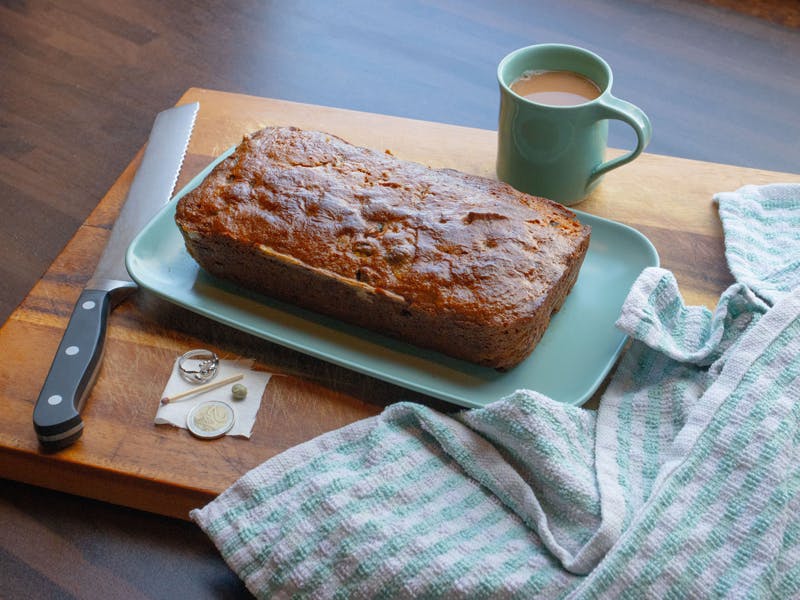 A freshly baked loaf of bread on a teal platter, accompanied by a cup of coffee, a striped kitchen towel, a serrated knife, and a few small items on a wooden cutting board.