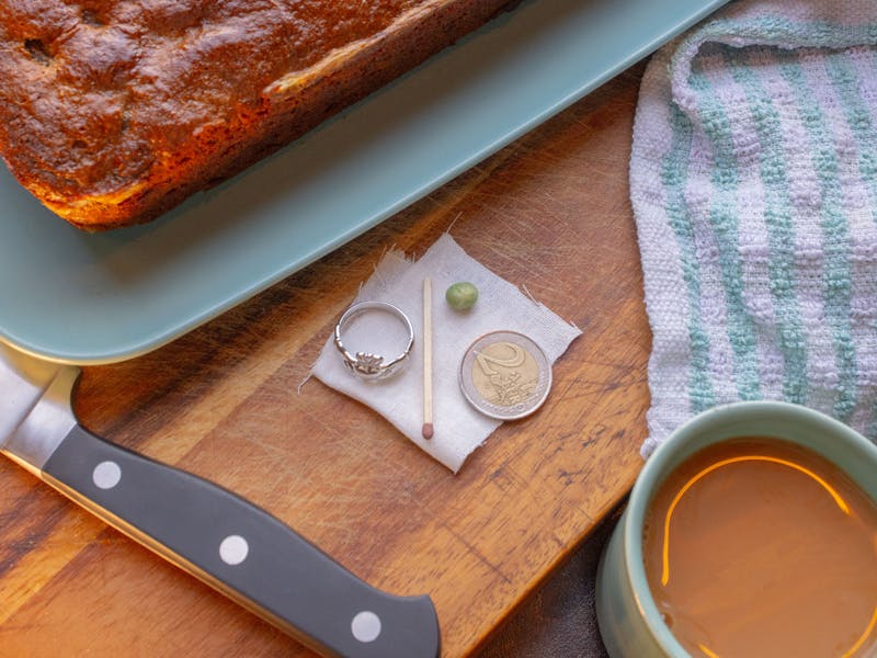 A loaf of bread on a teal serving plate next to a wooden cutting board displaying a ring, a matchstick, a green pea, and a coin with a coffee mug in the corner.
