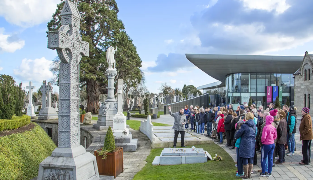 A tour guide speaks to a large group of people gathered in a cemetery, featuring ornate gravestones and sculptures, with a modern building visible in the background under a blue sky.