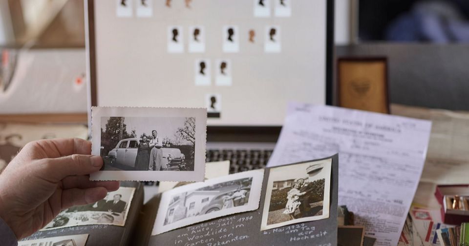 A person holding a vintage black and white photograph in front of an open scrapbook filled with old photos and documents, alongside a laptop displaying a family tree chart.
