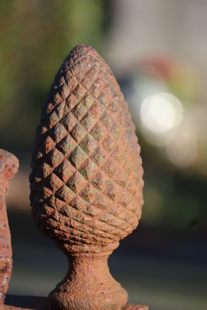 Close-up of a weathered iron pinecone finial with a textured surface, set against a blurred background.