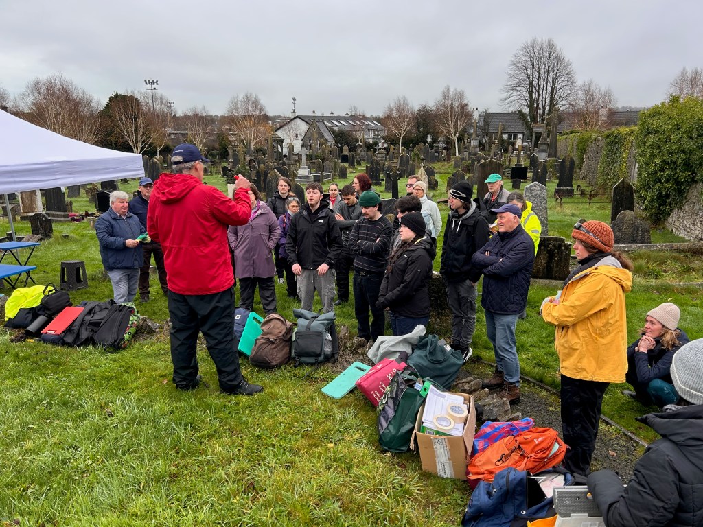A group of people gathered in a cemetery listening to a speaker in a red jacket, with gravestones visible in the background.