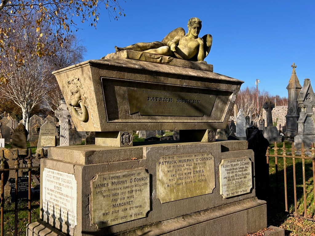 A monumental grave marker featuring a reclining angel statue atop a stone tomb, surrounded by various other gravestones and a clear blue sky.