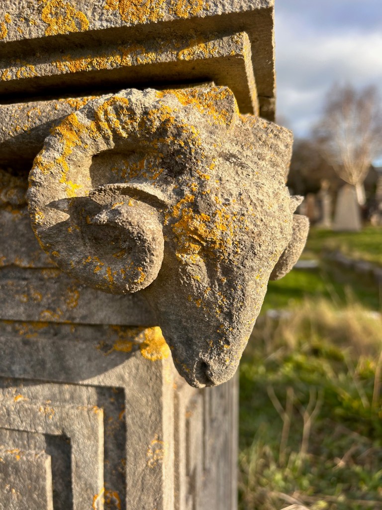 Close-up of a weathered stone ram sculpture with moss and lichen growth, situated on a pillar in a grassy outdoor setting.