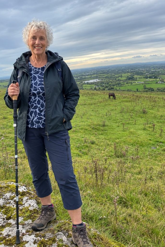 A smiling woman with curly hair stands on a grassy hill, holding a hiking pole, with a scenic landscape of fields and a river in the background.