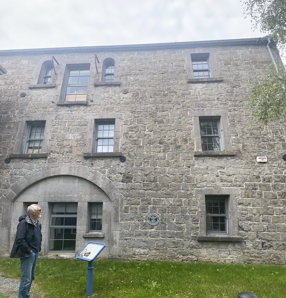 A man standing in front of a historic stone building with several windows and an arched entrance, accompanied by an informational sign.