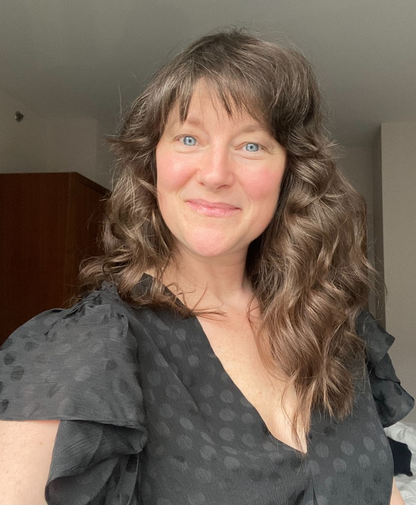 A person with wavy brown hair poses for a selfie, wearing a black polka dot top with ruffled sleeves, smiling in a well-lit indoor setting.