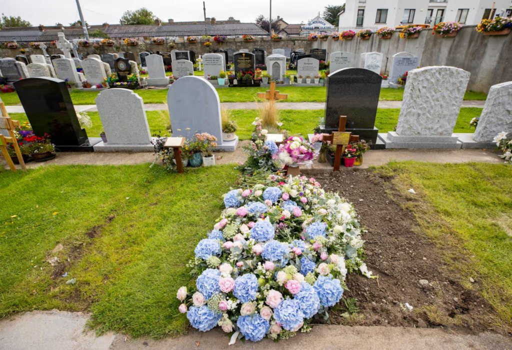 A cemetery scene with various gravestones, featuring a recently prepared grave adorned with blue and pink floral arrangements.