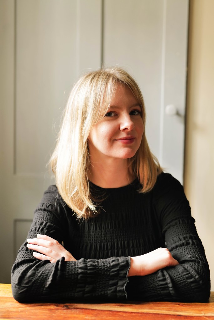A woman with long blonde hair smiles while sitting at a wooden table, wearing a black long-sleeve top. She has her arms crossed and is positioned in front of a light-colored background.