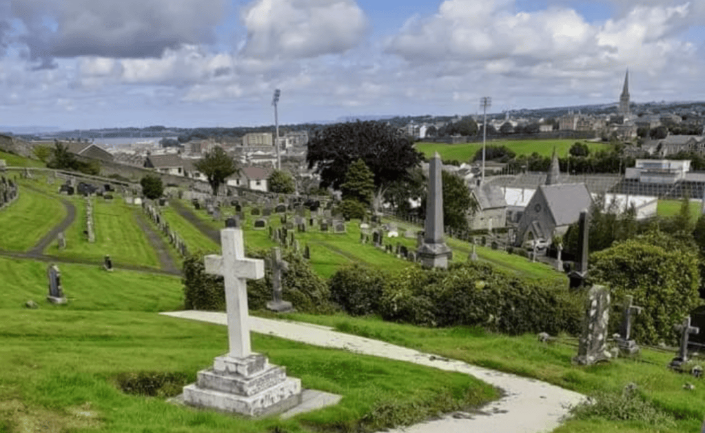 A scenic view of a cemetery on a hillside, featuring gravestones and monuments, with a backdrop of a town and sky filled with clouds.